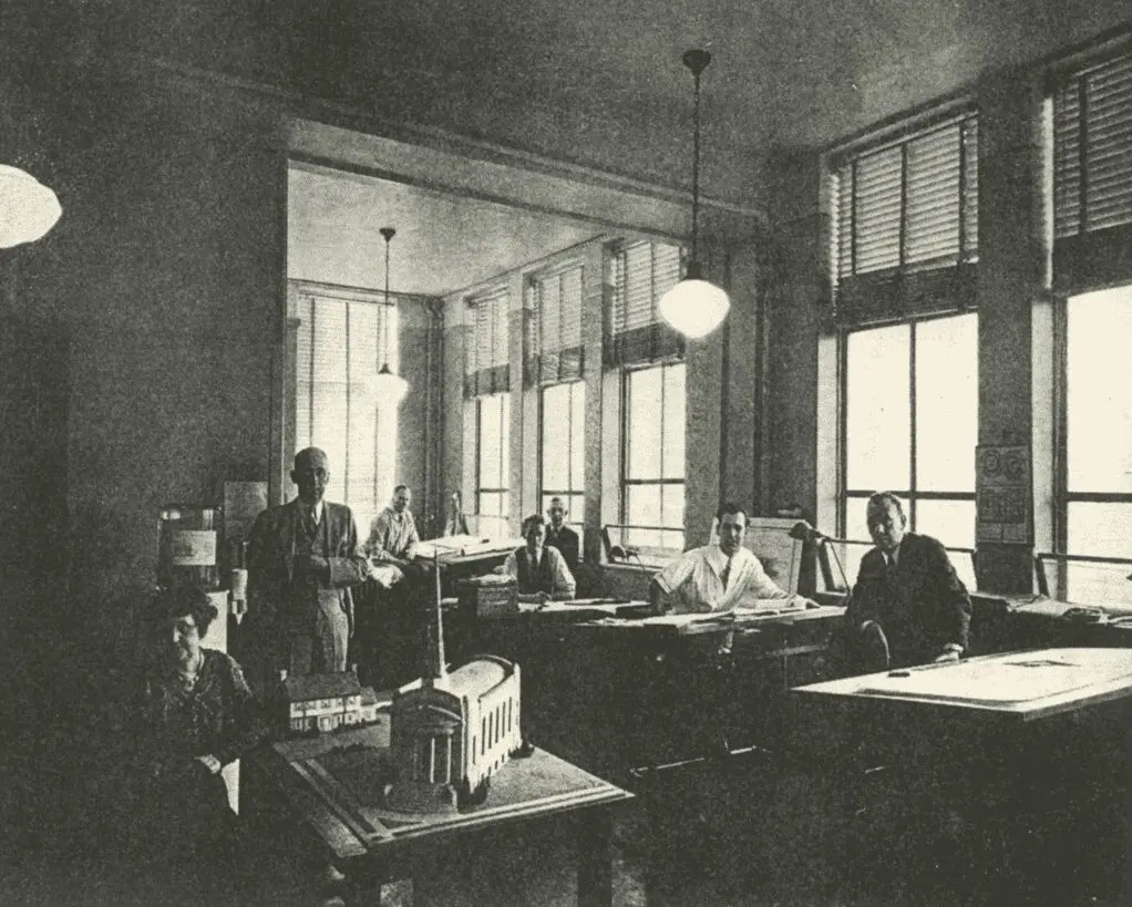 MS-office-1-jpg A group of seven people work at drafting tables in a well-lit room with large windows and overhead lights. Architectural models and blueprints are visible throughout the space.